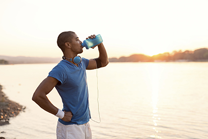 Cropped shot of a young sportsman drinking water while running on riverbank. Warm sunset tones.